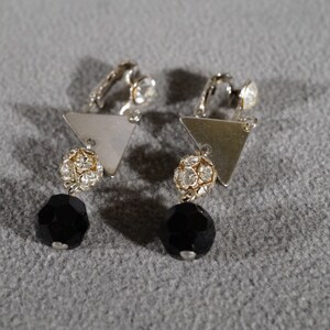 May include: A pair of silver clip-on earrings with black and clear crystal accents. The earrings feature a triangular shape with a dangling black crystal bead.