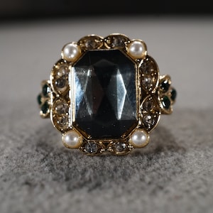 May include: A gold ring with a large, dark gray gemstone surrounded by smaller clear and white gemstones. The ring has green accents on the sides.