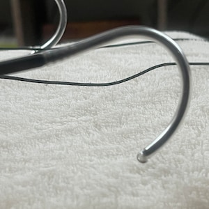 May include: Close-up of a silver medical device with a curved, hook-like end. The device is lying on a white, fluffy surface.