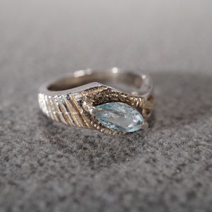 May include: A silver-toned ring with a marquise-cut, light blue gemstone. The ring features a textured band with gold-toned accents around the gemstone setting. The ring is a piece of jewelry.