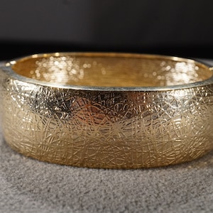 Puede incluir: Un brazalete dorado con una superficie texturizada y un patrón.