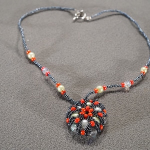 May include: A black beaded necklace with a pendant made of orange and white beads. The pendant is a round, flower-like design.