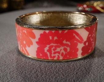 May include: A gold bangle bracelet with a pink and orange floral design. The bracelet is made of metal and has a smooth finish.
