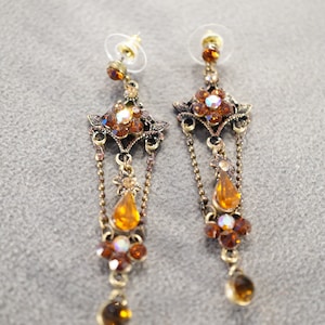 May include: A pair of long, dangling earrings with a gold tone finish. The earrings feature multiple tiers of amber-colored crystals and a teardrop-shaped crystal at the bottom.