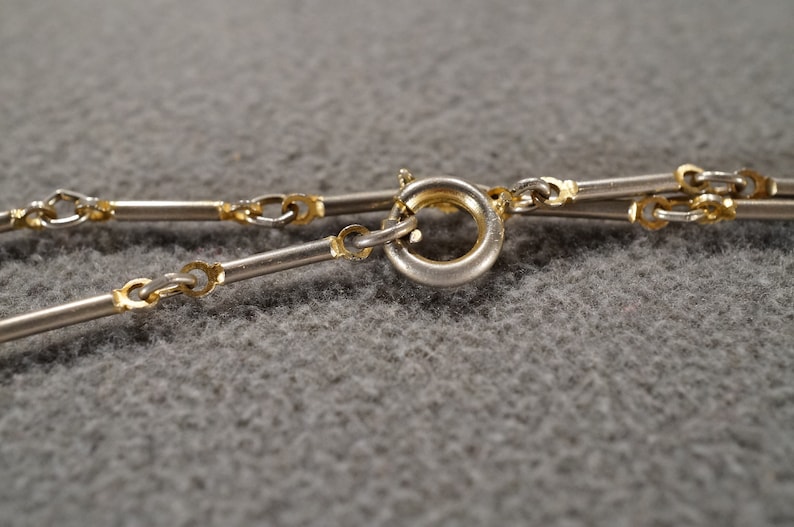 May include: Close-up of a silver and gold chain necklace with a clasp. The chain is made of small, linked circles and rectangular bars.