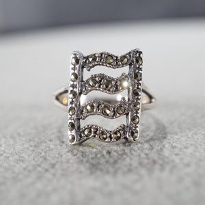 May include: A silver-toned ring with a rectangular design. The ring features a series of horizontal, wavy lines, each adorned with small, dark gemstones. The ring's band is simple and polished, creating a contrast with the detailed centerpiece.