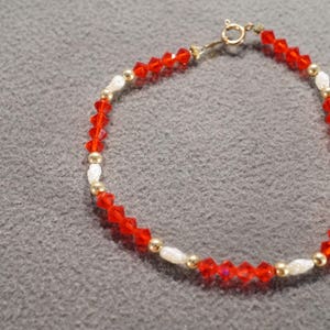 May include: A delicate bracelet featuring alternating red faceted beads, small gold-colored beads, and white oval beads. The bracelet has a gold-colored clasp. The jewelry is displayed on a gray surface.