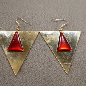May include: A pair of gold triangle earrings with red gemstone accents. The earrings have a geometric design and are a modern style.