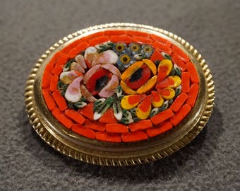 Peut inclure: Une broche ronde dorée avec un motif en mosaïque représentant des fleurs rouges, jaunes, roses et vertes. La mosaïque est encadrée d'un cadre circulaire avec un bord texturé.