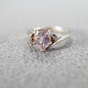 May include: A silver ring with a pear-shaped, light purple gemstone as the centerpiece. Two smaller, round gemstones flank the main stone. The ring is set against a gray background.