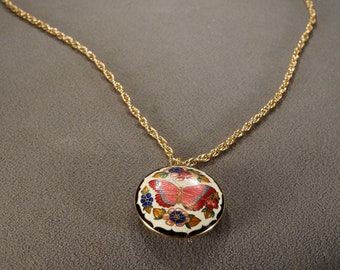 May include: A gold chain necklace with a round pendant featuring a hand-painted butterfly and floral design. The pendant is white with black outlining and colorful accents.