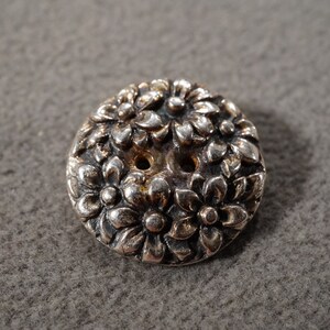 May include: A silver-toned button with a floral design. The button is round and features a raised pattern of flowers and leaves. It has two holes for sewing and is likely made of metal.