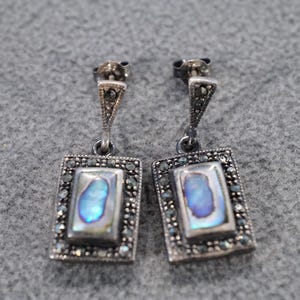 May include: A pair of silver-toned drop earrings. Each earring features a rectangular abalone shell center, framed by small, dark stones. The earrings have a decorative triangular top with additional small stones.
