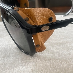 May include: Black aviator sunglasses with brown leather side shields. The sunglasses have a metal logo that says "Wings" on the side.