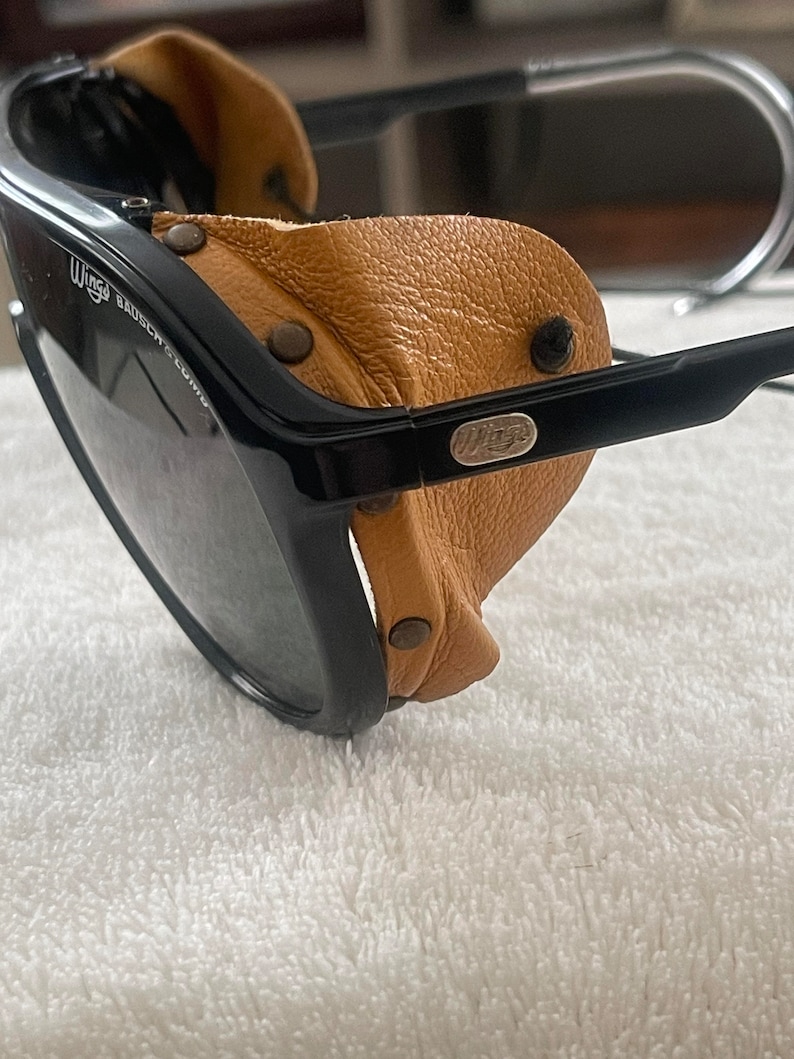 May include: Black aviator sunglasses with brown leather side shields. The sunglasses have a metal logo that says "Wings" on the side.