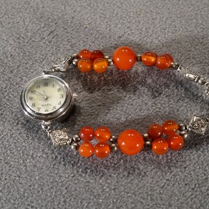 May include: A silver watch with a white face and orange beads attached to the band. The watch face says "QUARTZ".