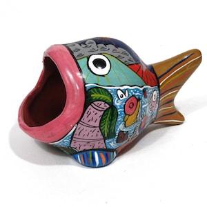 May include: A colorful, hand-painted ceramic fish-shaped container. The fish has a wide-open mouth with a pink rim, a large eye, and a tail. The body is decorated with various scenes and colors, including blue, green, and red.
