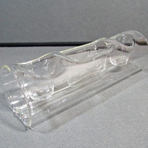 May include: A clear glass vase with a unique, curved design. The vase has a cylindrical shape with a wavy, decorative element running along its length. The glass appears to be smooth and transparent, allowing light to pass through.