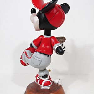 May include: A Mickey Mouse figurine in a baseball uniform. The figure wears a red baseball cap, shirt, and shoes, with grey trousers. The figurine is on a brown base with a baseball and a bat.