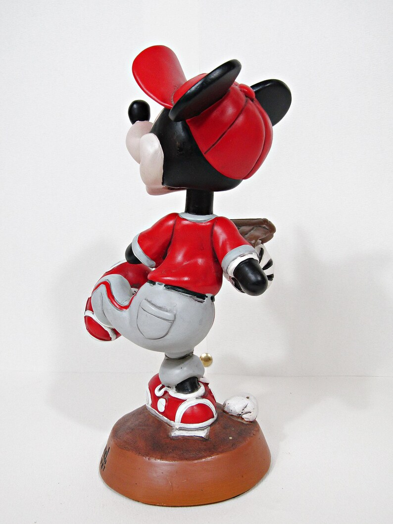 May include: A Mickey Mouse figurine in a baseball uniform. The figure wears a red baseball cap, shirt, and shoes, with grey trousers. The figurine is on a brown base with a baseball and a bat.