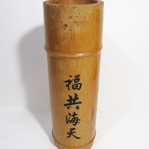 Puede incluir: Un jarrón de bambú cilíndrico y alto de color marrón natural. El jarrón tiene caracteres chinos negros inscritos en el lateral que se leen "福寿海天".