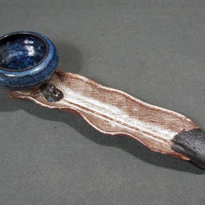 May include: A ceramic spoon with a deep, glossy, dark blue circular bowl and a long, textured, wavy handle. The handle features a brown, weathered finish with a white-washed center and a solid black tip at the end of the elongated stem.