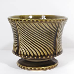 May include: A decorative ceramic planter in shades of olive green and brown. The planter features a flared shape with a textured, diagonal stripe pattern and a pedestal base. The rim and base are adorned with small, raised dots.
