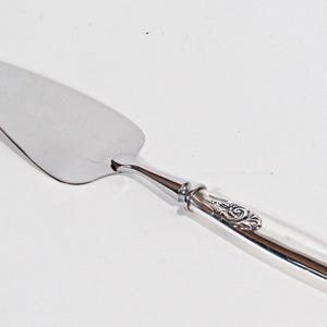 May include: A silver cake server with a polished blade and a decorative handle. The handle features an ornate design and a curved shape. This serving utensil is ideal for cakes and pies.