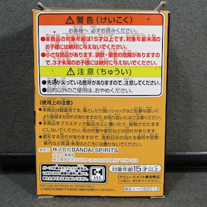 May include: A rectangular box with Japanese text in orange, yellow, and white. The text includes warnings and age restrictions. The box also features the Bandai Spirits logo and a product code.
