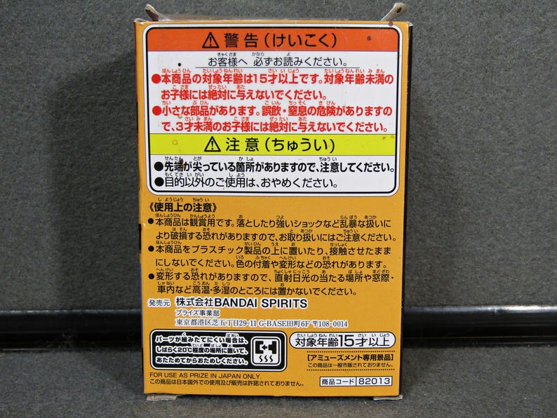 May include: A rectangular box with Japanese text in orange, yellow, and white. The text includes warnings and age restrictions. The box also features the Bandai Spirits logo and a product code.