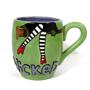 Puede incluir: Taza de cerámica verde con diseño de bruja. La taza presenta las piernas de una bruja con medias a rayas blancas y negras y zapatos rojos, con la palabra "Wicked" en letras grises. El asa es verde y el interior es morado.
