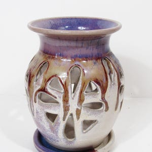 May include: A ceramic vase with a round body and a flared rim. The vase is white with cut-out designs and is adorned with purple and brown glaze. It sits on a matching circular base. The vase is suitable for home decor.
