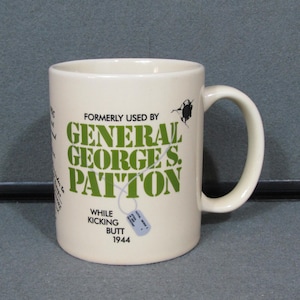 May include: Cream-colored ceramic mug with a handle. The mug features green text that reads "FORMERLY USED BY GENERAL GEORGE S. PATTON" and "WHILE KICKING BUTT 1944". A small dog tag graphic is also present.