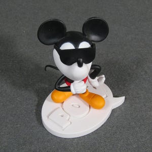 May include: A Mickey Mouse figurine wearing sunglasses, a red bow tie, and orange shoes. The figure is posed on a white base with the word "POP" in relief. The figure's ears are black, and the face is white.