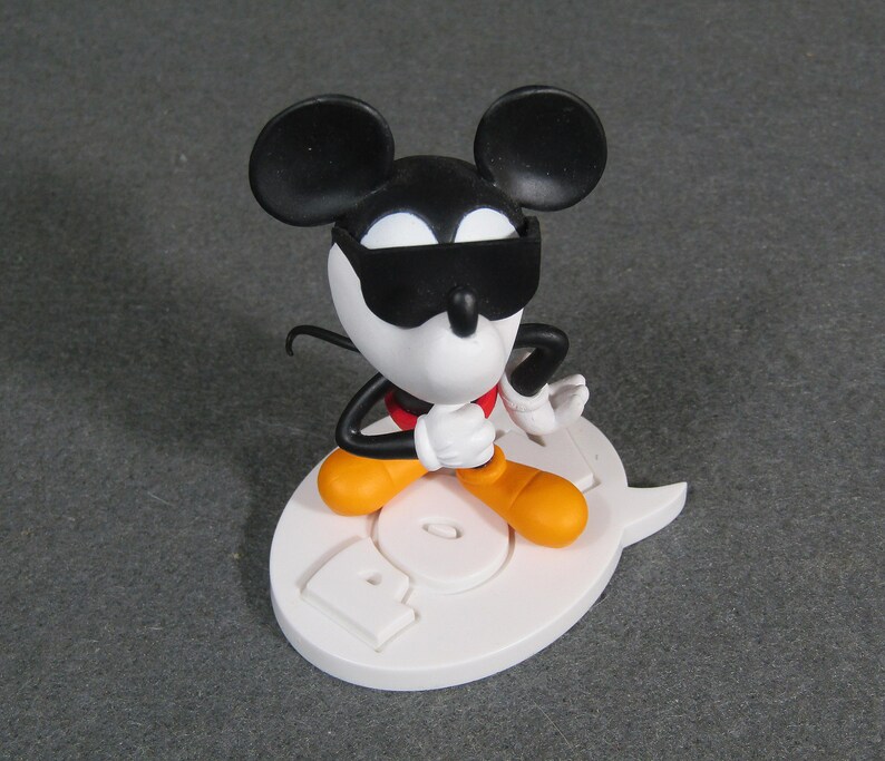 May include: A Mickey Mouse figurine wearing sunglasses, a red bow tie, and orange shoes. The figure is posed on a white base with the word "POP" in relief. The figure's ears are black, and the face is white.