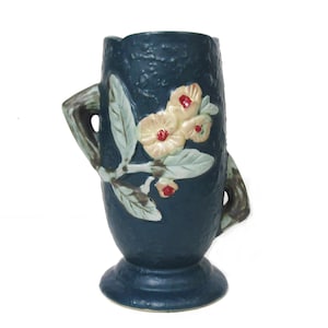 May include: A dark teal ceramic vase with a textured surface and a flared base. The vase features a floral design with cream-colored flowers, green leaves, and red accents. It has two curved handles.