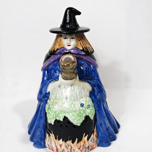 May include: A ceramic witch-shaped teapot with a black hat, blonde hair, and a blue robe adorned with stars. The teapot's base is a cauldron with flames, and the spout is a textured, cylindrical shape. The witch's face is painted with delicate features.