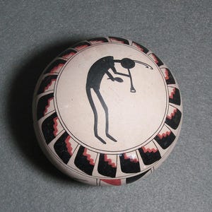 May include: A round, off-white ceramic pot with a black and red geometric border. The center features a black silhouette of a flute player. The pot has a smooth, rounded shape and is likely handmade.