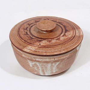 May include: A round, lidded ceramic bowl in shades of brown and white. The lid features a decorative pattern and a small knob. The bowl's exterior has a speckled appearance, suggesting a handmade quality. The design is suitable for kitchen or dining use.