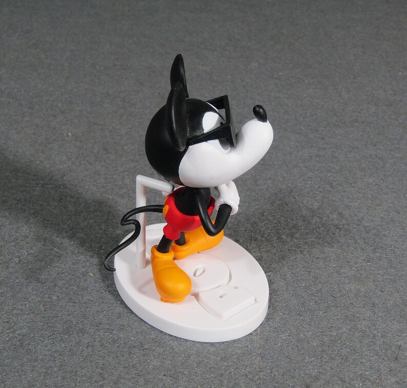 May include: A figurine of Mickey Mouse wearing sunglasses, red shorts, yellow shoes, and white gloves. The black and white figure is posed on a white base with a white support. The figurine is a collectible item.