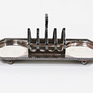 May include: A silver-colored toast rack with a handle and four slots, set on a tray with two round compartments. The tray has a decorative beaded edge and rests on small ball feet. A classic piece for serving toast.