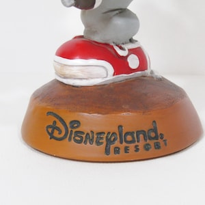 May include: A Disneyland Resort figurine featuring a grey character wearing a red shoe with white accents. The base is brown with the Disneyland Resort logo in black.