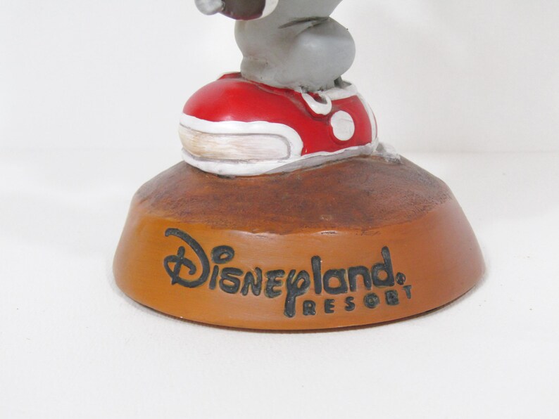 May include: A Disneyland Resort figurine featuring a grey character wearing a red shoe with white accents. The base is brown with the Disneyland Resort logo in black.
