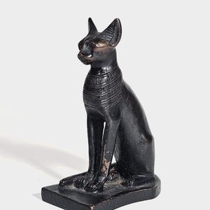 Puede incluir: Estatua de gato negro en posición sentada, con orejas erguidas y un collar detallado. La estatua está sobre una base rectangular y tiene una apariencia desgastada, lo que sugiere una pieza antigua o vintage. Los rasgos del gato están bien definidos.