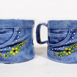 May include: Two blue ceramic mugs designed to resemble denim jeans. Each mug features a handle, a pocket design, and is decorated with yellow and green floral accents. The mugs are set against a white background.