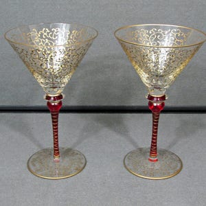 May include: Two clear martini glasses with gold filigree designs on the bowl. The stems are a deep red with a spiral design, and the base is clear with a gold rim. The glasses are elegant and decorative.