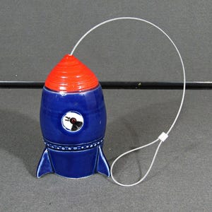 May include: A ceramic rocket-shaped bottle stopper. The rocket has a blue body, a red top, and blue fins. A white wire loop is attached to the top. The rocket has a small, round, white and black graphic on the front.
