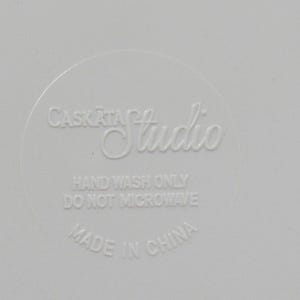 May include: A close-up of a white surface with the embossed text "Caskata Studio". The text "Hand Wash Only" and "Do Not Microwave" are also visible. The words "Made in China" are at the bottom.