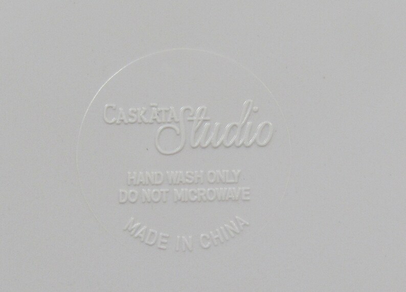 May include: A close-up of a white surface with the embossed text "Caskata Studio". The text "Hand Wash Only" and "Do Not Microwave" are also visible. The words "Made in China" are at the bottom.