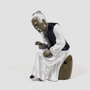 Op de afbeelding: Een keramiek beeldje van een oudere persoon die op een bruin vat zit. Het beeldje is geschilderd in tinten wit, bruin, blauw en grijs. De persoon draagt een witte robe en een blauw vest.